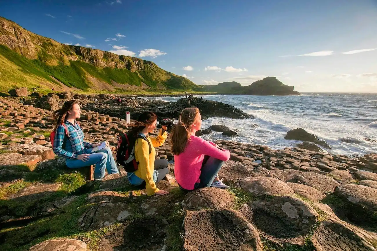 Three women sitting on the hexagonal basalt columns of the Giant's Causeway, looking out over the Atlantic Ocean.