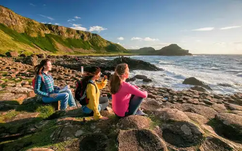 Three women sitting on the hexagonal basalt columns of the Giant's Causeway, looking out over the Atlantic Ocean.
