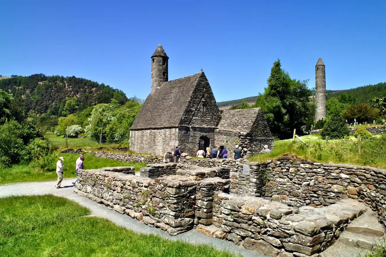 Esplora le storiche rovine monastiche del VI secolo e la famosa torre rotonda di Glendalough. Antiche rovine monastiche in pietra che presentano una struttura di tetto tradizionale e una torre rotonda di pietra che si eleva sotto un cielo azzurro chiaro.