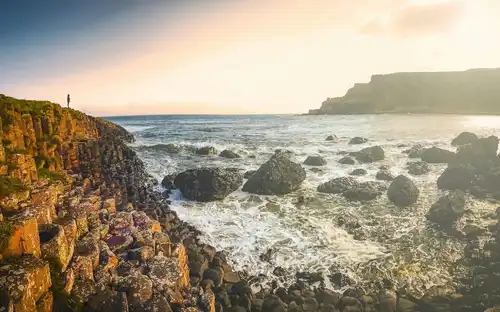 People walking along the dramatic hexagonal basalt columns of the Giant's Causeway with large green cliffs in the background.