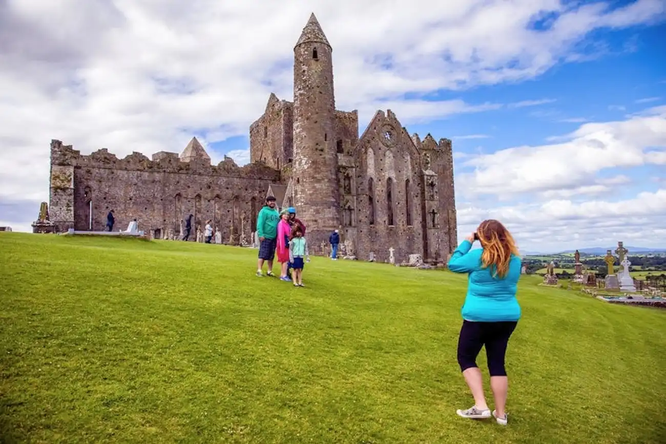 Tourists photographing the historic Rock of Cashel buildings perched on a green hill.