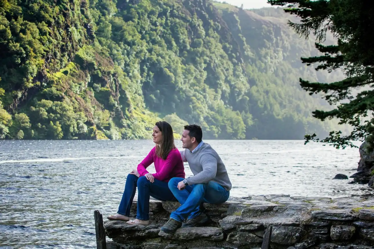 Goditi oltre 2 ore complete di tempo libero per rilassarti accanto ai laghi glaciali di Glendalough. Una coppia seduta insieme su un muro di pietra lungo le rive di un lago cristallino circondato da colline boscose.