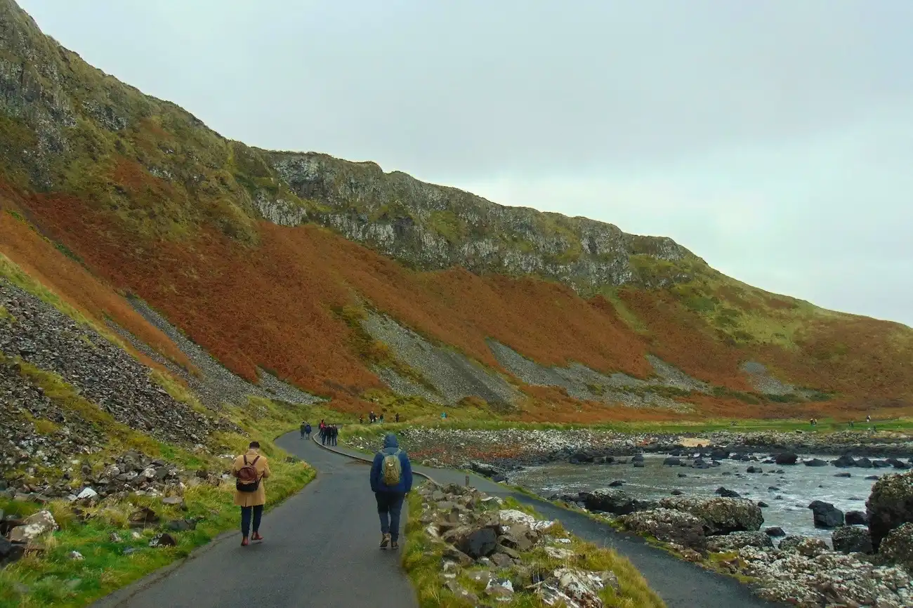Tourists walking along a paved coastal path towards the Giant's Causeway with large green cliffs in the background.