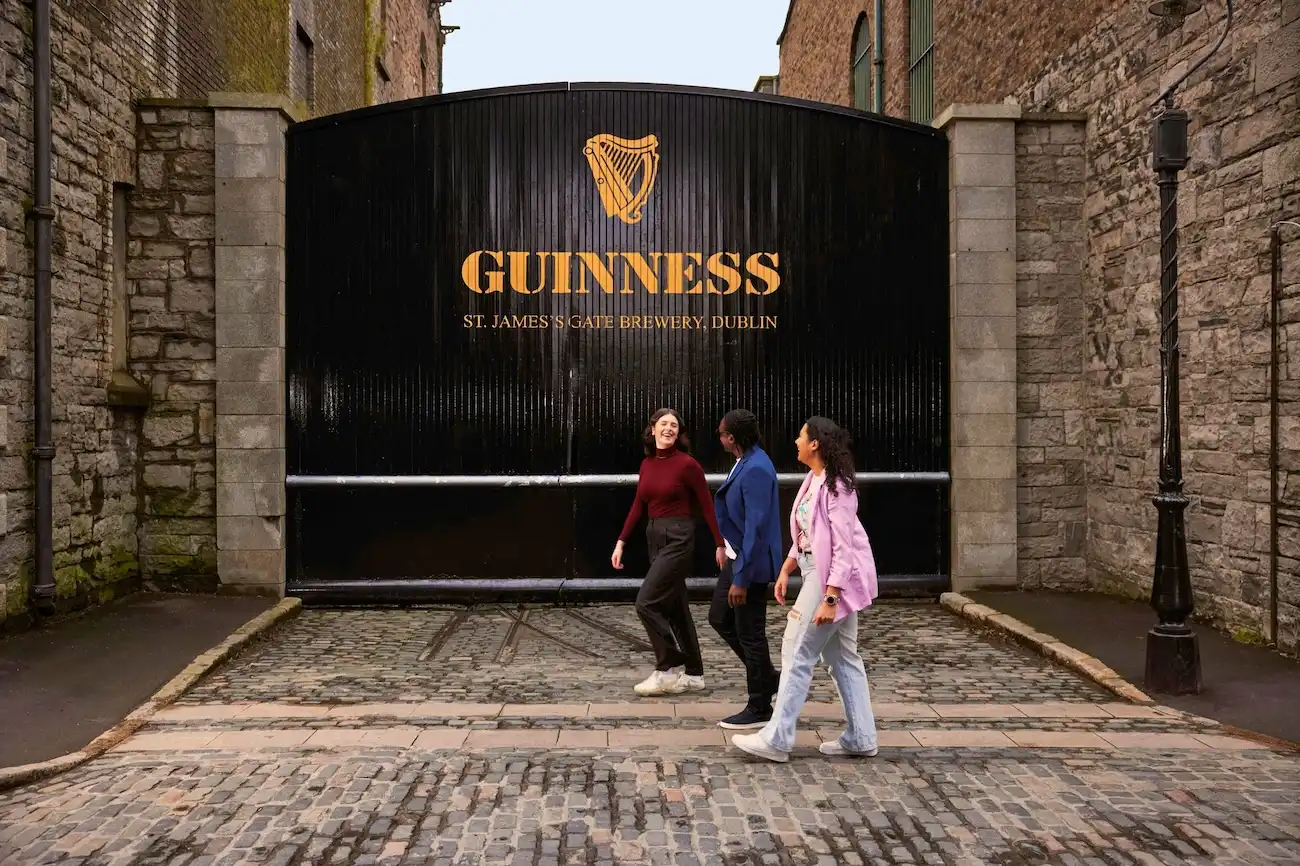 Three smiling friends walking in front of the iconic black and gold Guinness St. James's Gate.