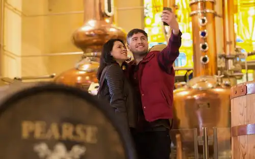 A happy couple taking a selfie inside Pearse Lyons Distillery with copper stills in the background.